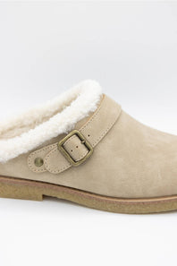 A close-up of a beige suede clog-style slipper with a white sherpa lining and an antique bronze buckle strap, shown against a plain white background.