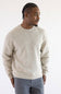 A man wearing a light beige knit crewneck sweater and gray trousers, standing against a plain white studio background.