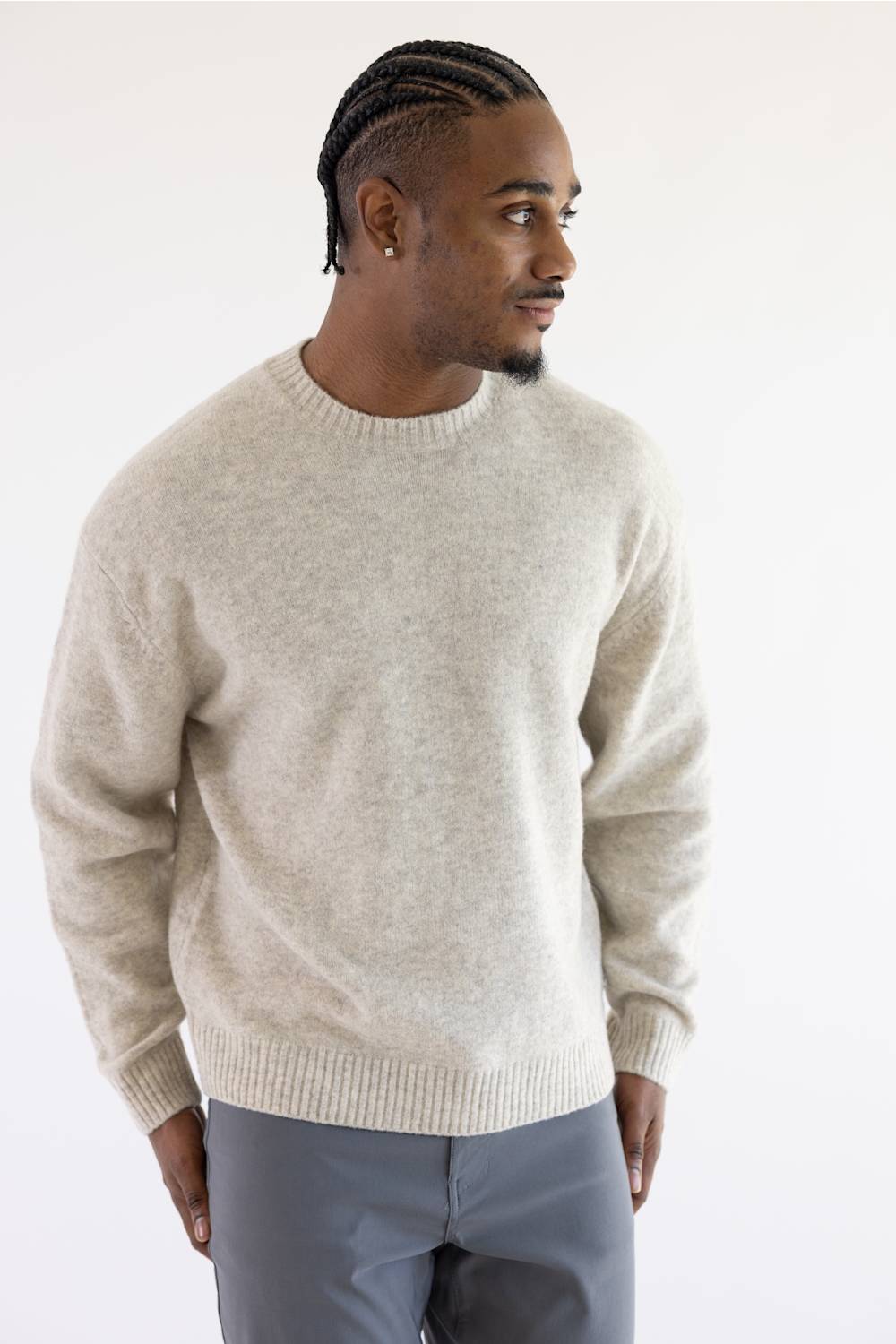 A man wearing a light beige knit crewneck sweater and gray trousers, standing against a plain white studio background.