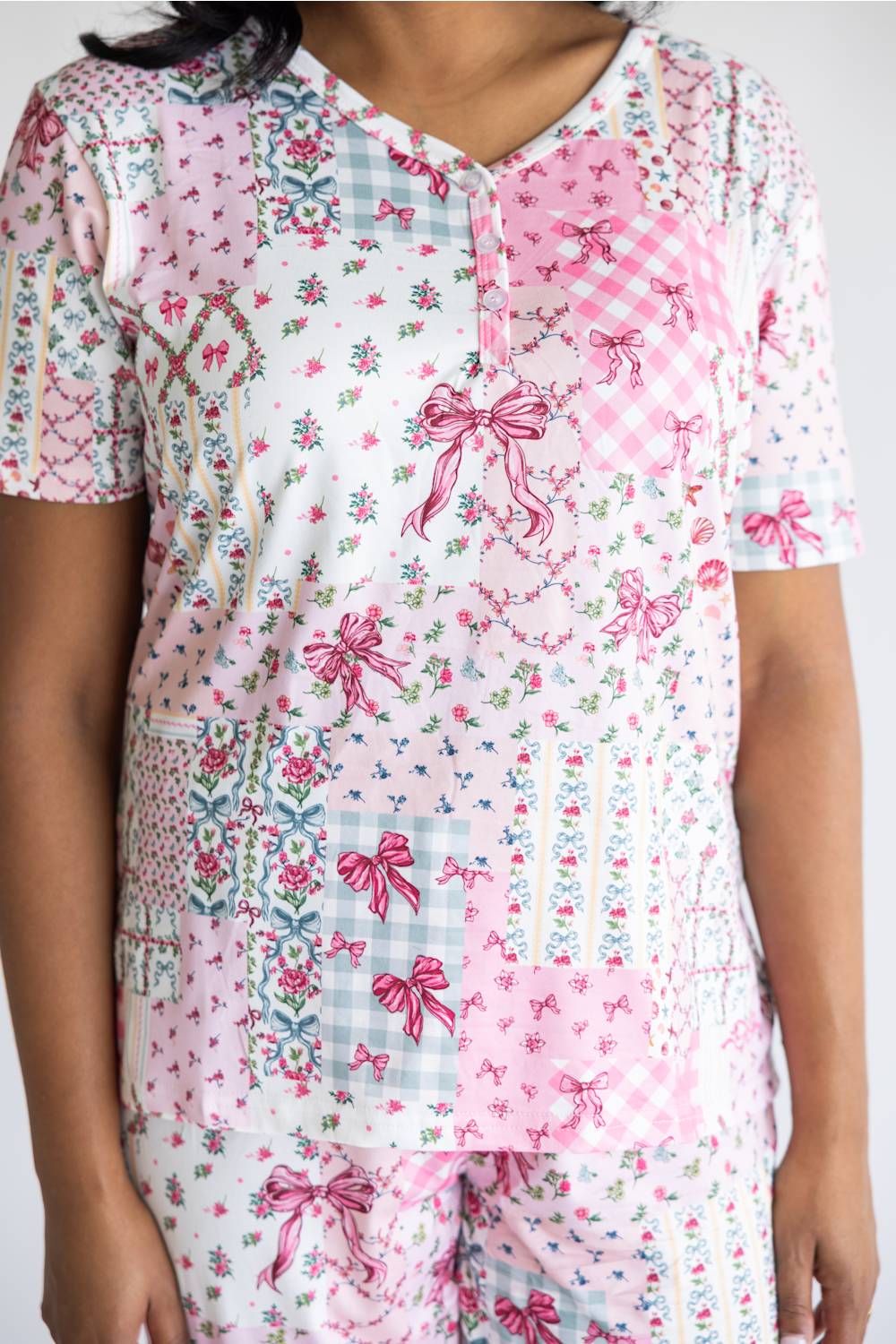 A woman wearing a short-sleeve V-neck pajama top with a patchwork print of pink gingham, pink bows, floral motifs, and light blue gingham panels, paired with matching pajama pants against a plain light background.