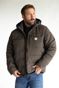 A man wearing a dark brown Carhartt hooded puffer jacket with a black shirt and blue jeans, standing against a light gray background.