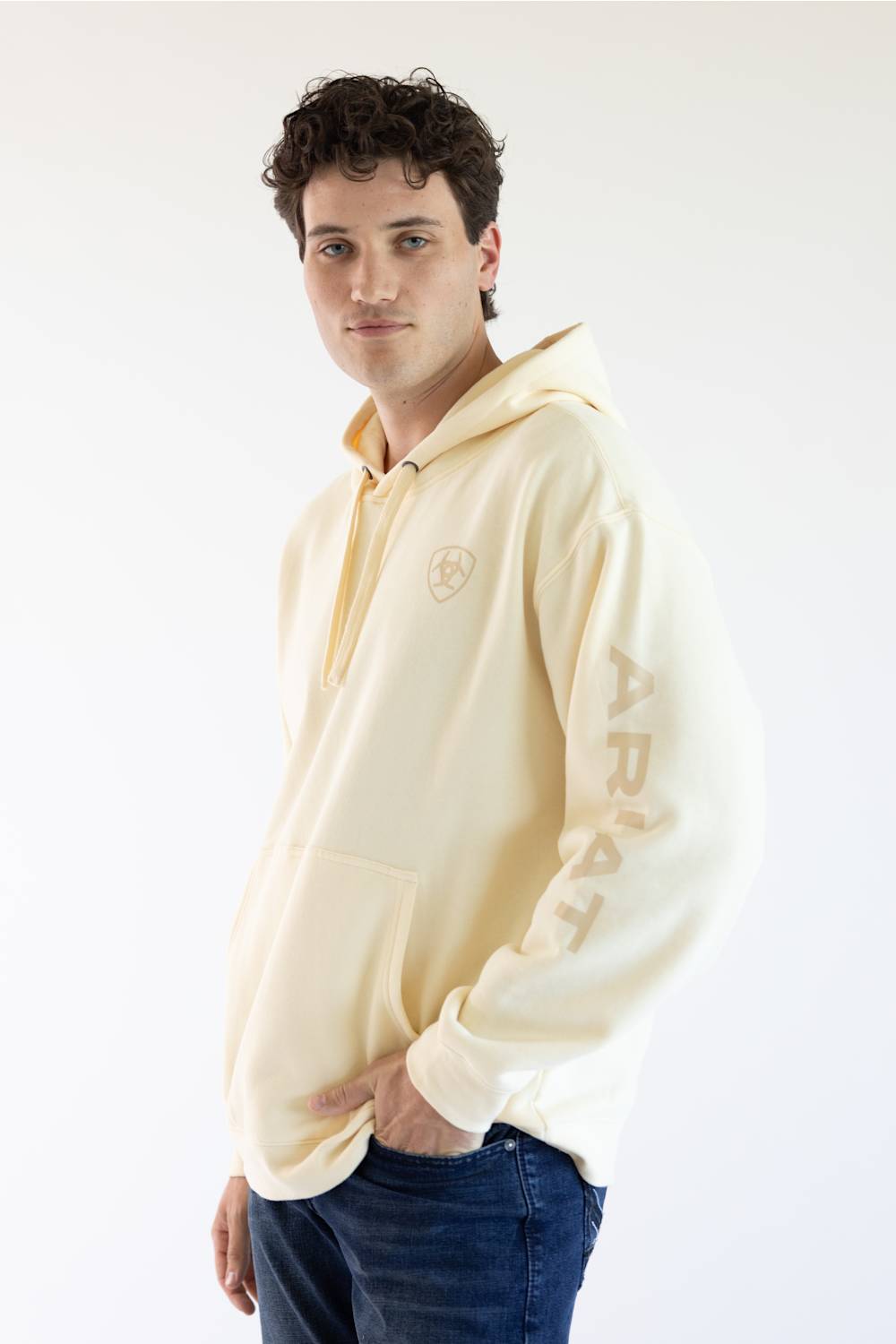 A man wearing a cream-colored hoodie with a small circular logo on the left chest and vertical lettering on the right sleeve, paired with blue jeans, photographed against a plain white studio background.