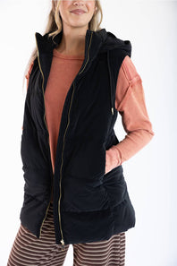 A woman wearing a black hooded puffer vest over a peach long-sleeve shirt and striped pants, photographed against a plain white studio background.