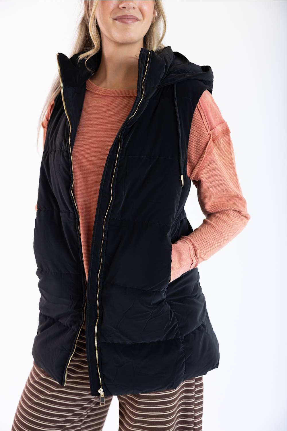 A woman wearing a black hooded puffer vest over a peach long-sleeve shirt and striped pants, photographed against a plain white studio background.