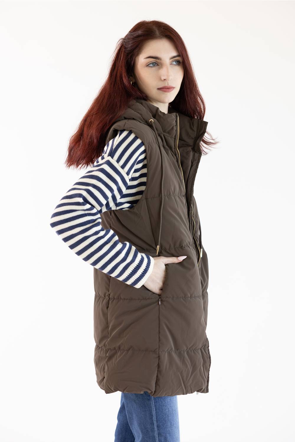 A woman with red hair models a dark brown puffer vest over a navy and white striped long-sleeve shirt and blue jeans, photographed against a plain white background.