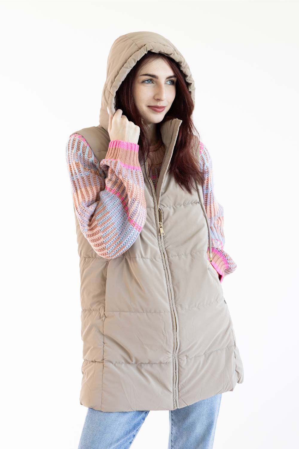 A woman wearing a beige hooded puffer vest over a pastel striped knit sweater and blue jeans, photographed against a plain white studio background.