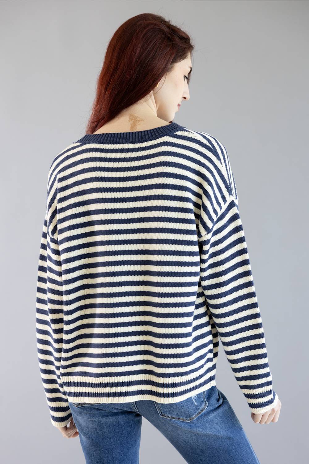 A woman shown from the back wearing a navy and cream striped knit sweater with blue jeans, set against a plain gray studio background.