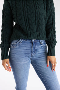 A person wearing a dark forest green cable-knit sweater and light blue jeans, shown from the chest down against a plain white background.