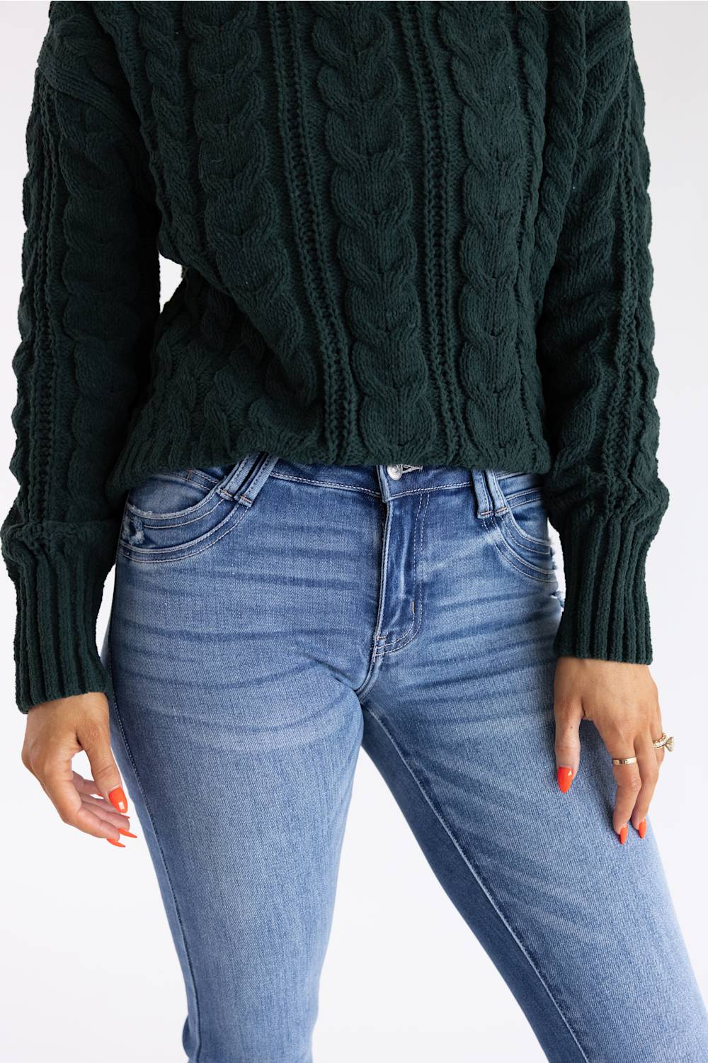 A person wearing a dark forest green cable-knit sweater and light blue jeans, shown from the chest down against a plain white background.