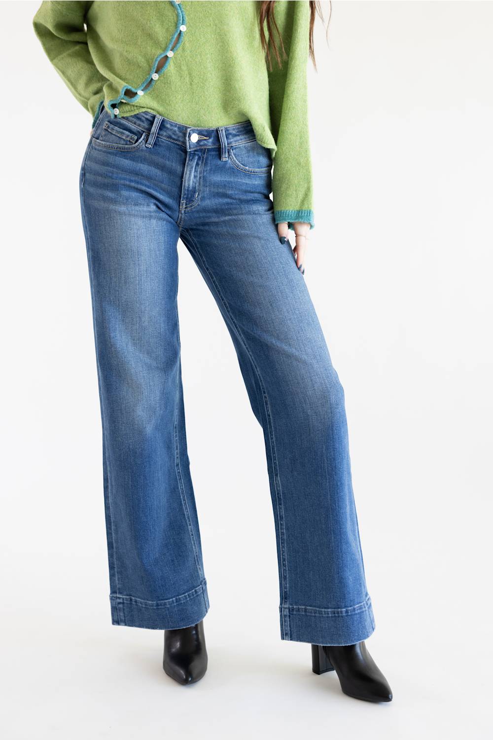 A person wearing blue wide-leg denim jeans, a light green sweater, and black ankle boots, photographed against a plain white studio background.