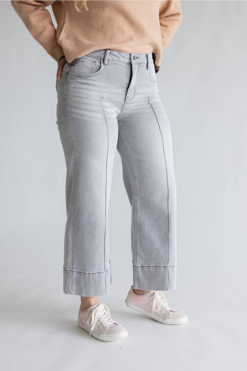 A person wearing light gray wide-leg cropped jeans with rolled cuffs, paired with a beige sweater and white sneakers, shown from the waist down against a light gray studio background.