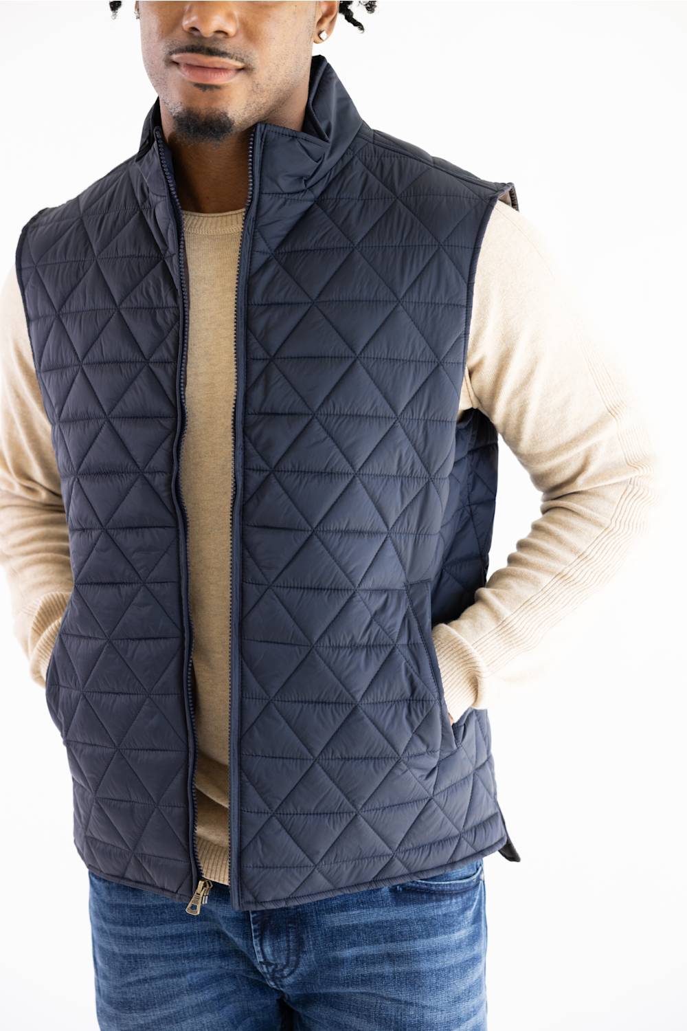 A man wearing a navy blue diamond-quilted zip-up puffer vest over a beige long-sleeve shirt and blue jeans, photographed against a plain white background.