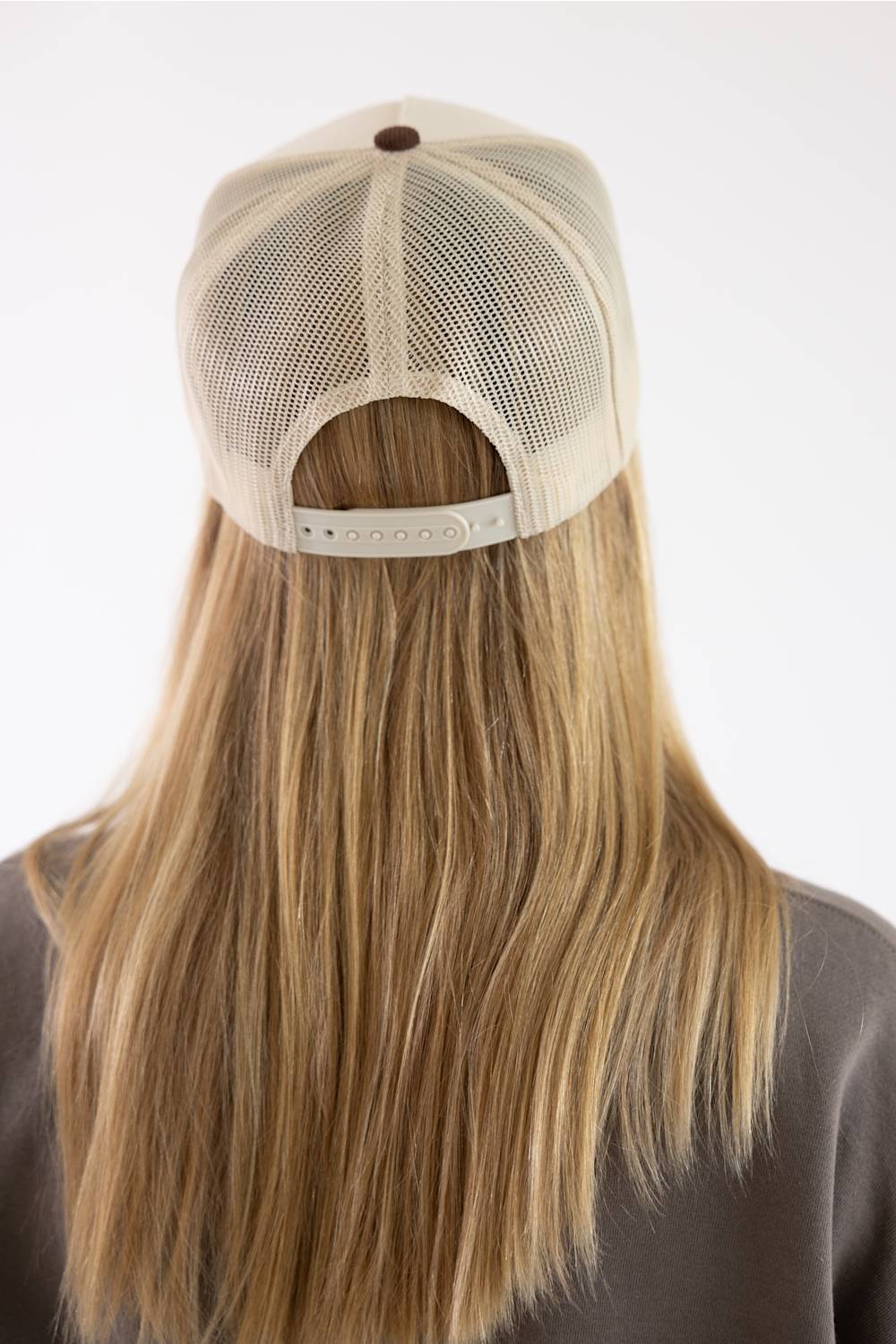 Back view of a person with long blonde hair wearing a beige mesh trucker cap with an adjustable strap, set against a plain white background.