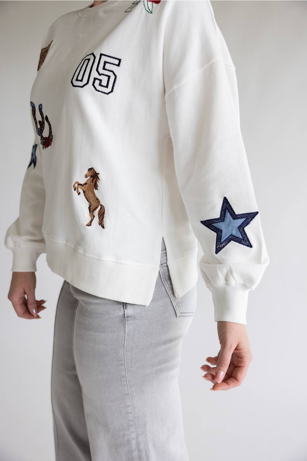 A person wearing a white crewneck sweatshirt with embroidered patches, including the number 05, a horse, and a blue star on the sleeve, paired with light gray pants.