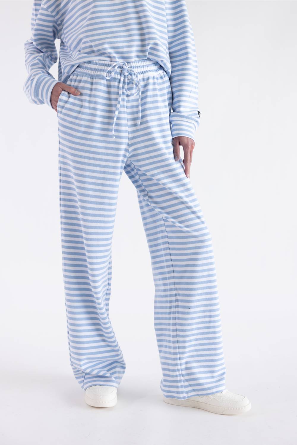 A person wearing light blue and white striped lounge pants with a drawstring waist and a matching striped long-sleeve top, paired with white slip-on shoes, against a plain light background.