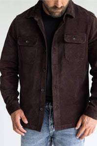 A man wearing a dark brown corduroy button-up shirt jacket over a black t-shirt and blue jeans, shown from the chin down against a light gray background.