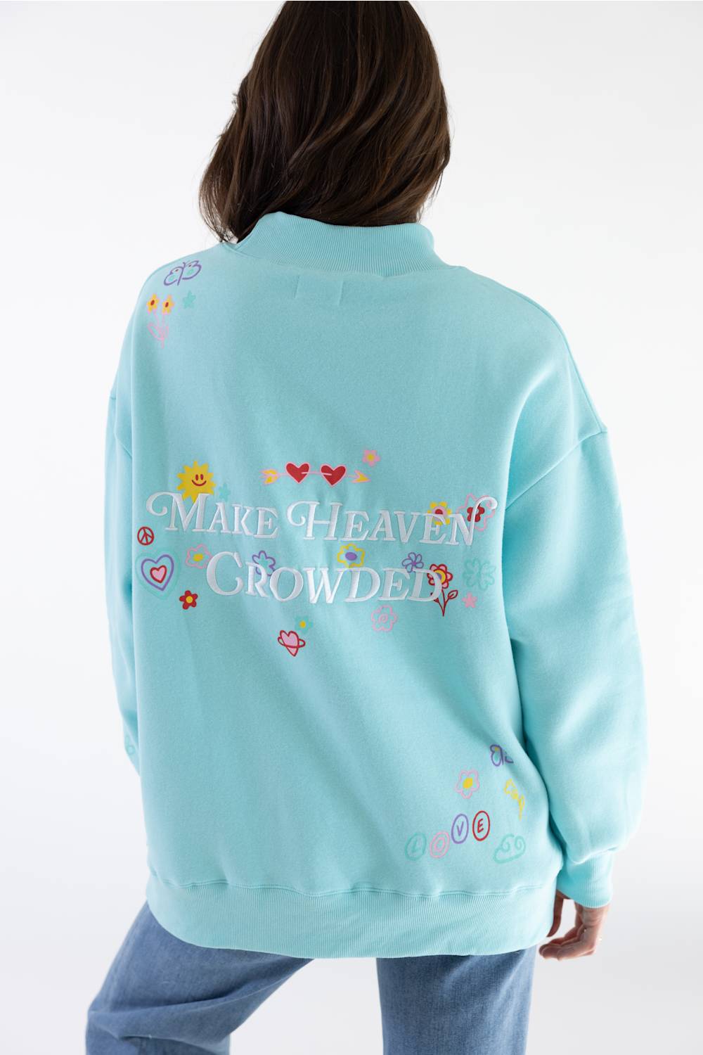 A person shown from the back wearing a light aqua blue sweatshirt with colorful embroidered graphics and the text 'MAKE HEAVEN CROWDed' across the back, paired with blue jeans against a plain white studio background.