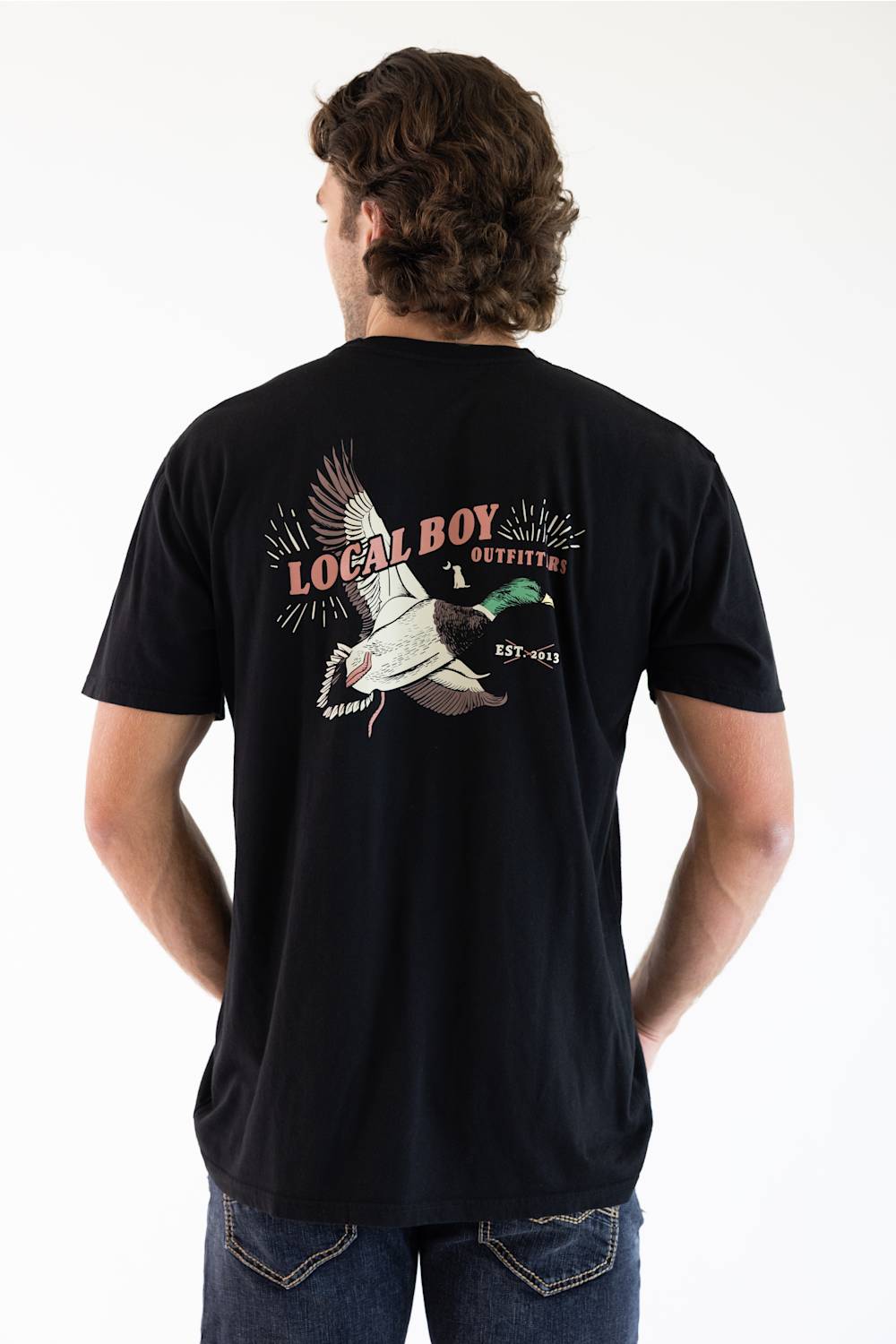 A man viewed from behind wearing a black t-shirt with a large graphic on the back featuring a mallard duck and the text 'LOCAL BOY OUTFITTERS EST. 2013'.