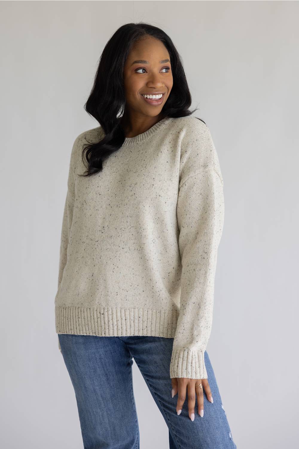 A woman wearing a cream-colored speckled knit sweater and blue jeans, standing against a plain light gray background.