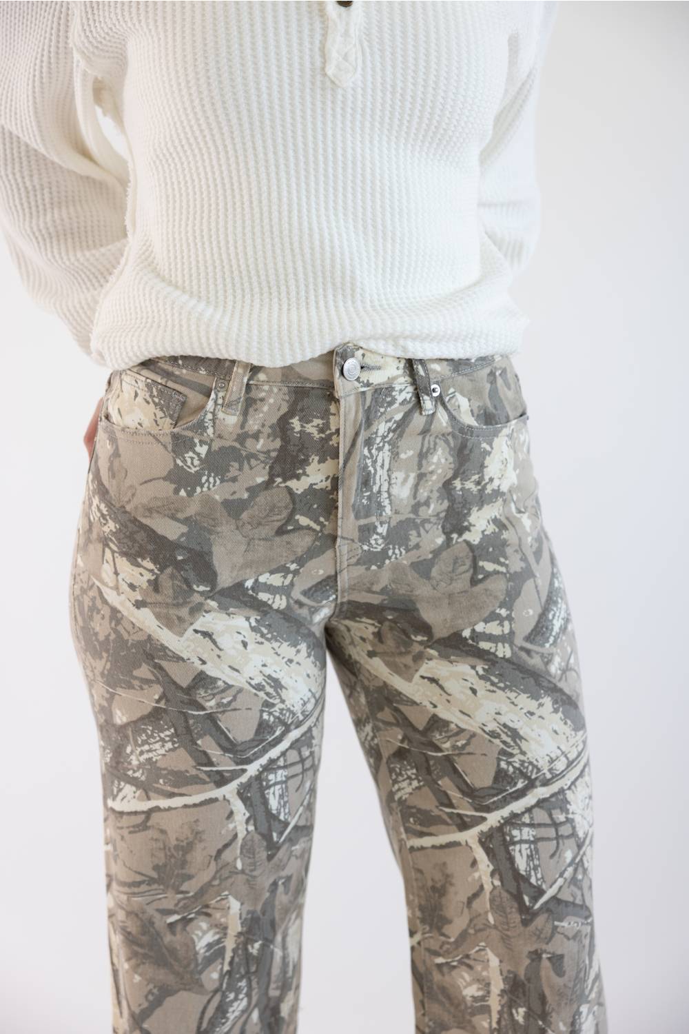 A person wearing camouflage-print pants and a white ribbed knit sweater, cropped from the chest down against a plain light background.