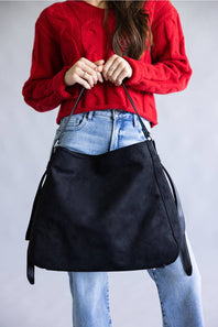 A person wearing a red cable-knit sweater and light blue jeans holds a large black suede hobo shoulder bag against a plain gray studio background.