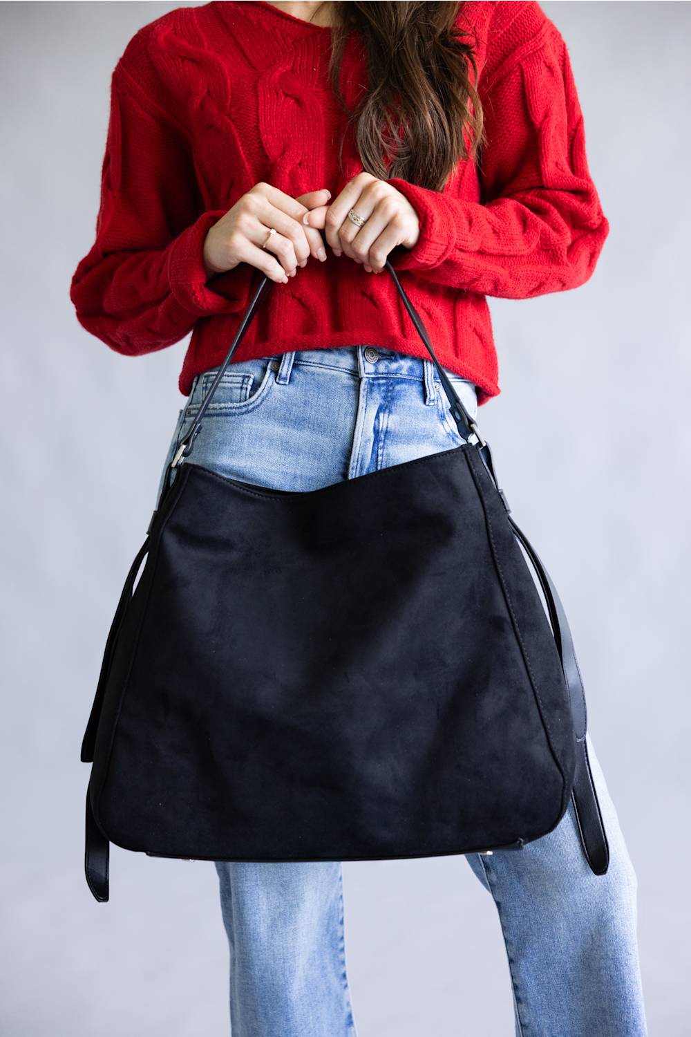 A person wearing a red cable-knit sweater and light blue jeans holds a large black suede hobo shoulder bag against a plain gray studio background.