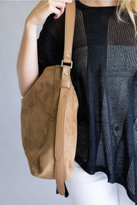 A tan suede shoulder bag with a matching adjustable strap and silver-tone hardware, worn by a model wearing a black mesh top and white pants against a light gray background.