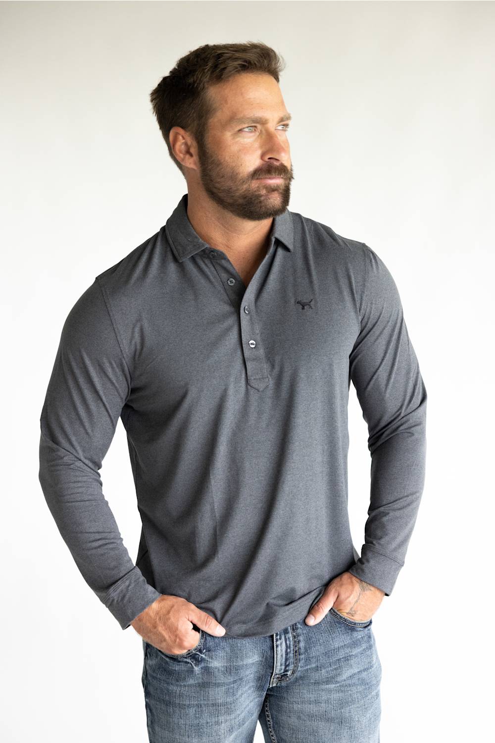 A muscular man wearing a dark gray long-sleeve polo shirt with a small embroidered logo on the left chest, paired with blue jeans, standing against a plain white studio background.