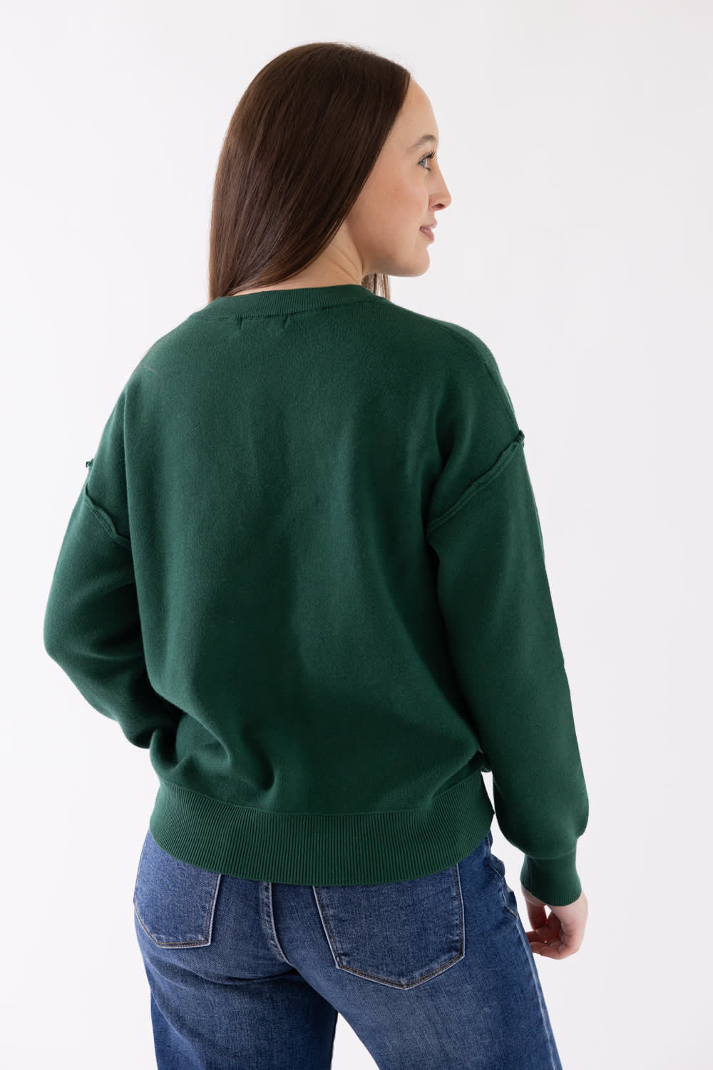 A woman wearing a dark green crewneck sweatshirt and blue jeans, shown from a side-back angle against a plain white studio background.