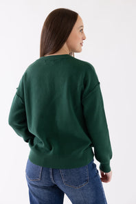 A woman wearing a dark green crewneck sweatshirt and blue jeans, shown from a side-back angle against a plain white studio background.