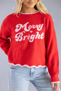 A woman wearing a bright red knit sweater with 'Merry Bright' written in white across the front, paired with light blue jeans, against a solid gray studio background.