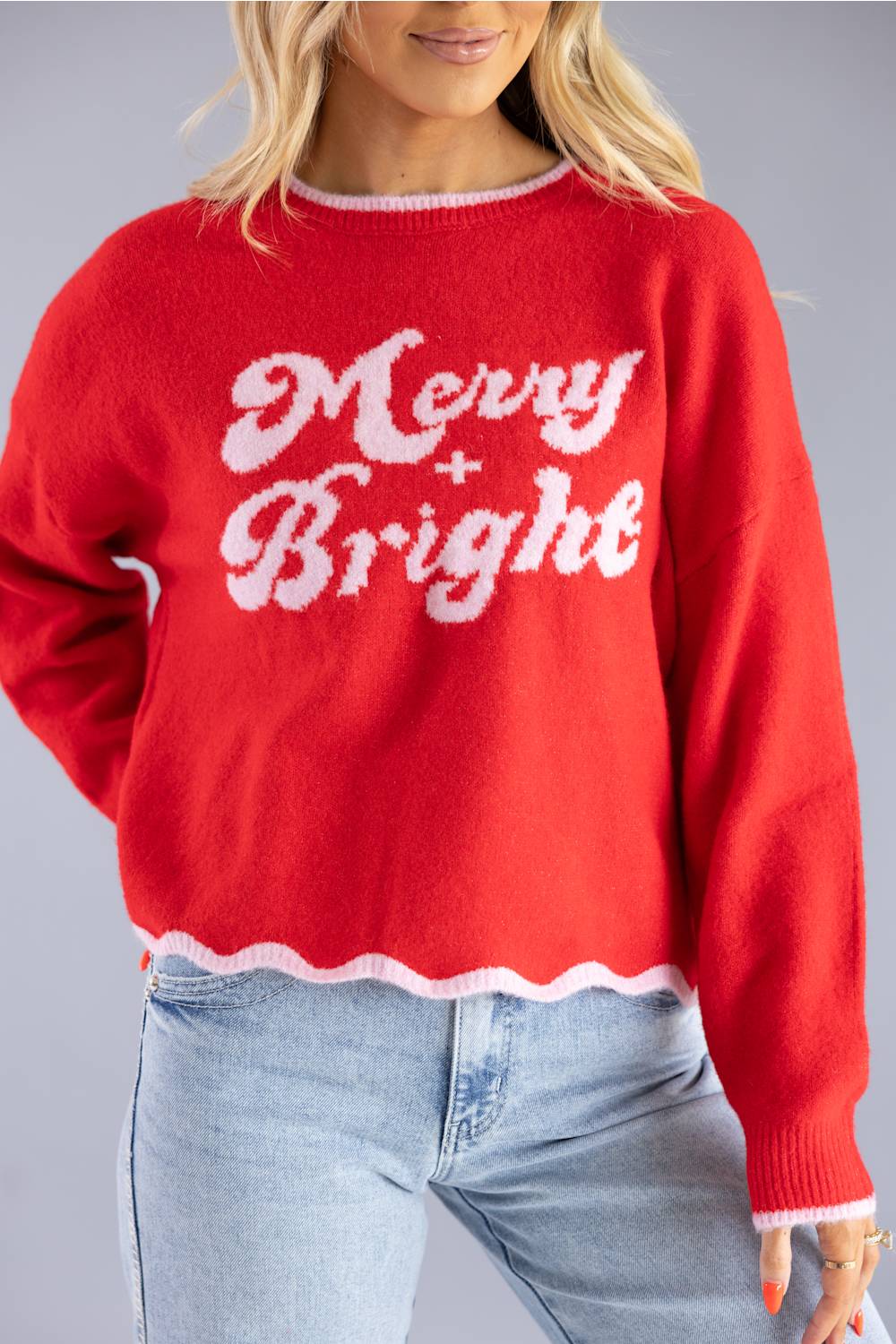 A woman wearing a bright red knit sweater with 'Merry Bright' written in white across the front, paired with light blue jeans, against a solid gray studio background.
