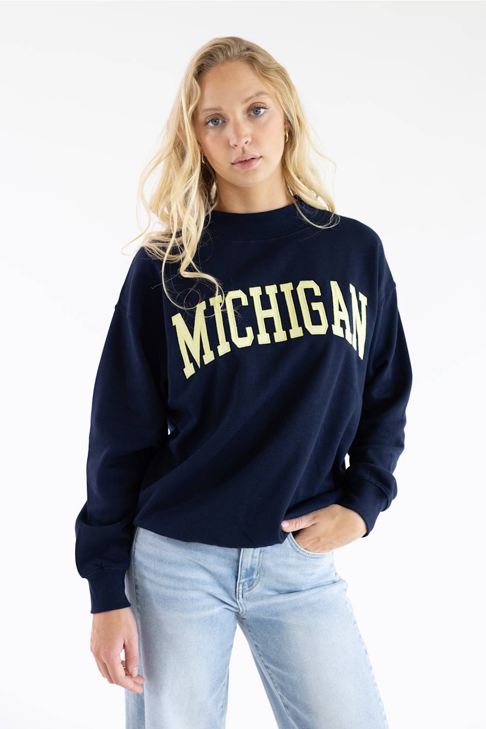 A blonde woman wearing a navy blue crewneck sweatshirt with 'MICHIGAN' printed in yellow across the chest, paired with light blue jeans, against a plain white background.