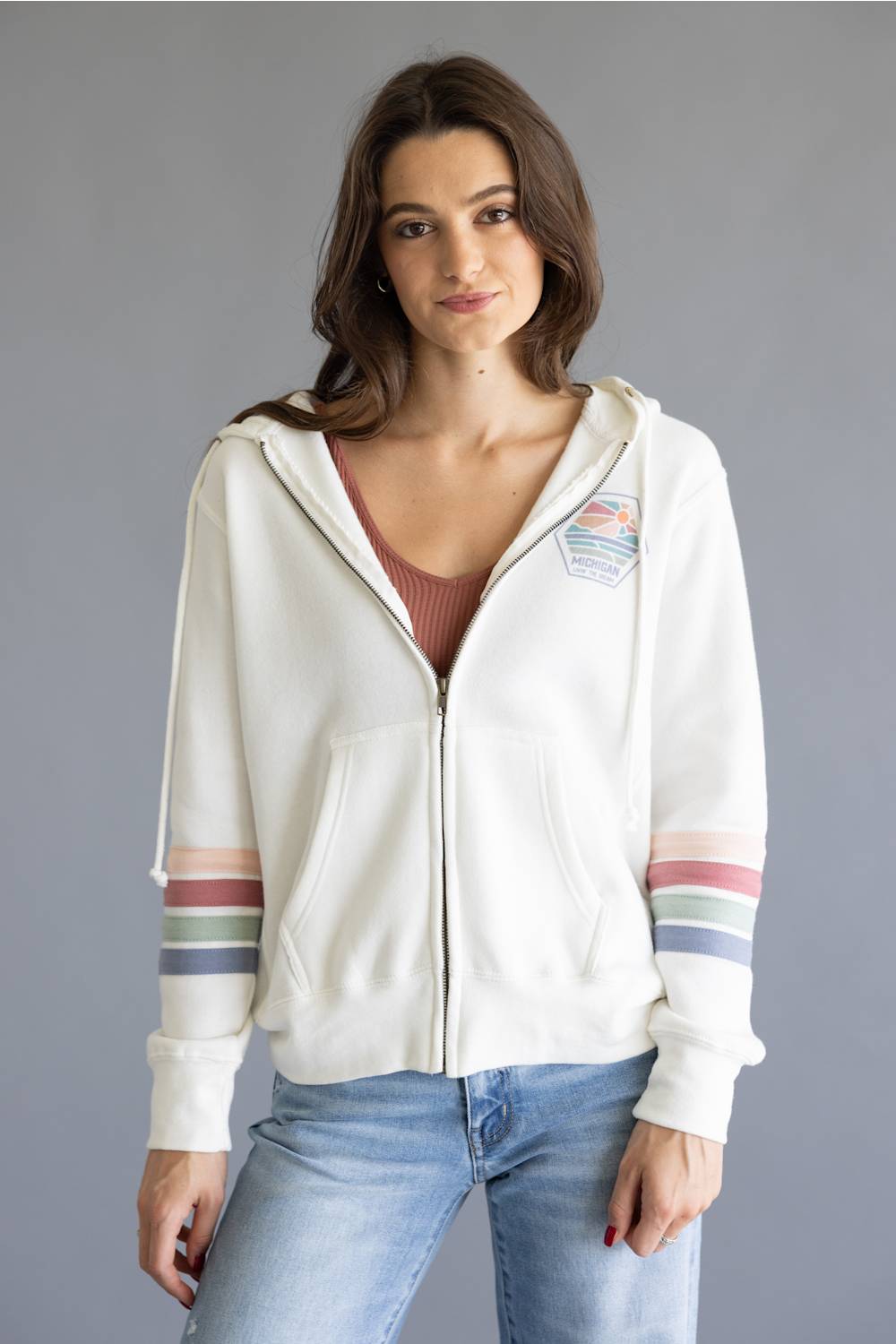 A woman wearing a white zip-up hoodie with colorful striped details on the sleeves and a small patch on the left chest, paired with light blue jeans, against a solid gray background.
