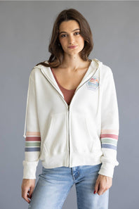 A woman wearing a white zip-up hoodie with colorful striped details on the sleeves and a small patch on the left chest, paired with light blue jeans, against a solid gray background.