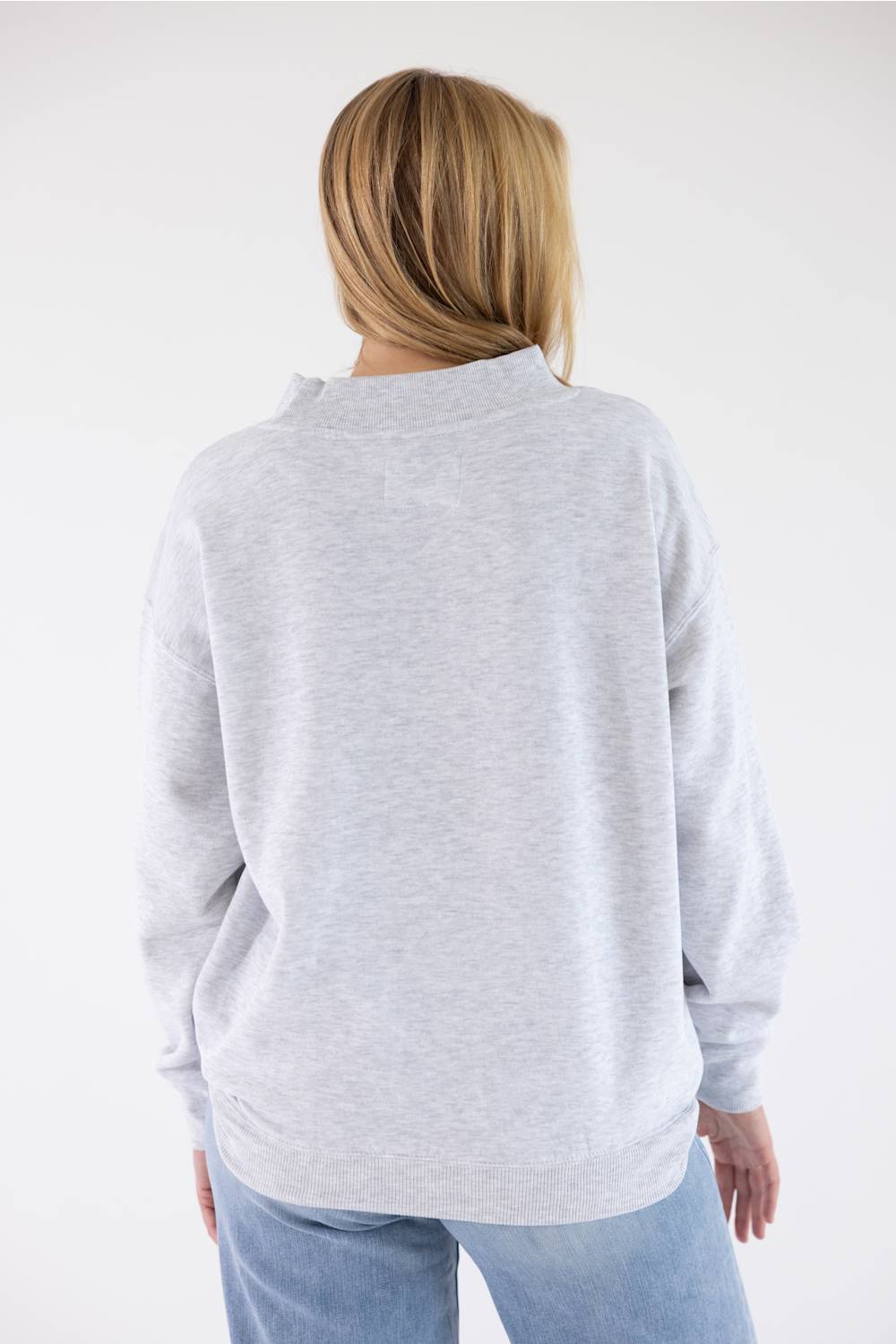 A woman viewed from behind wearing a light heather gray long-sleeve sweatshirt and light blue jeans against a plain white studio background.