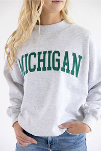 A woman wearing a light heather gray crewneck sweatshirt with 'MICHIGAN' printed in green varsity-style letters across the chest, paired with blue jeans.