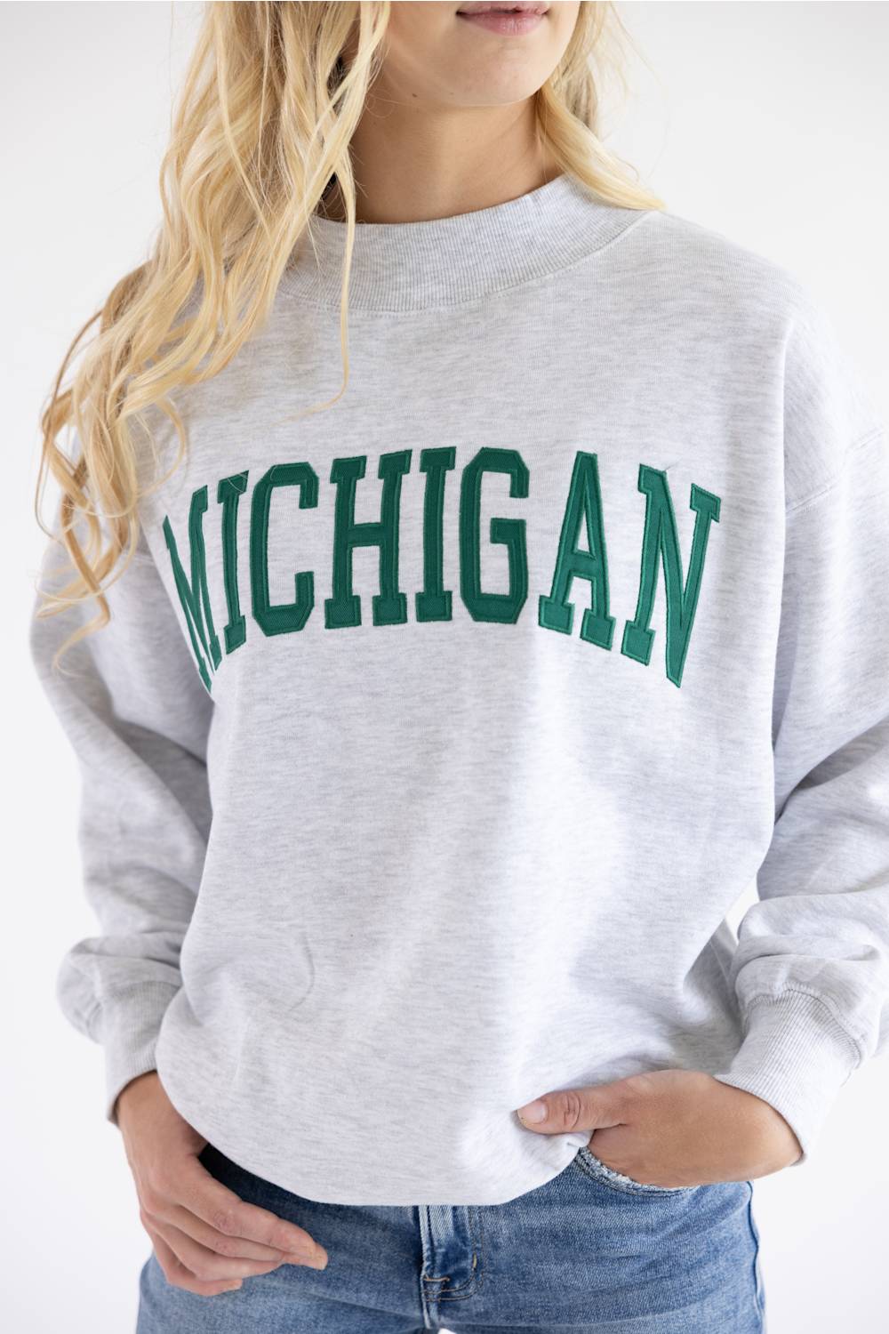 A woman wearing a light heather gray crewneck sweatshirt with 'MICHIGAN' printed in green varsity-style letters across the chest, paired with blue jeans.