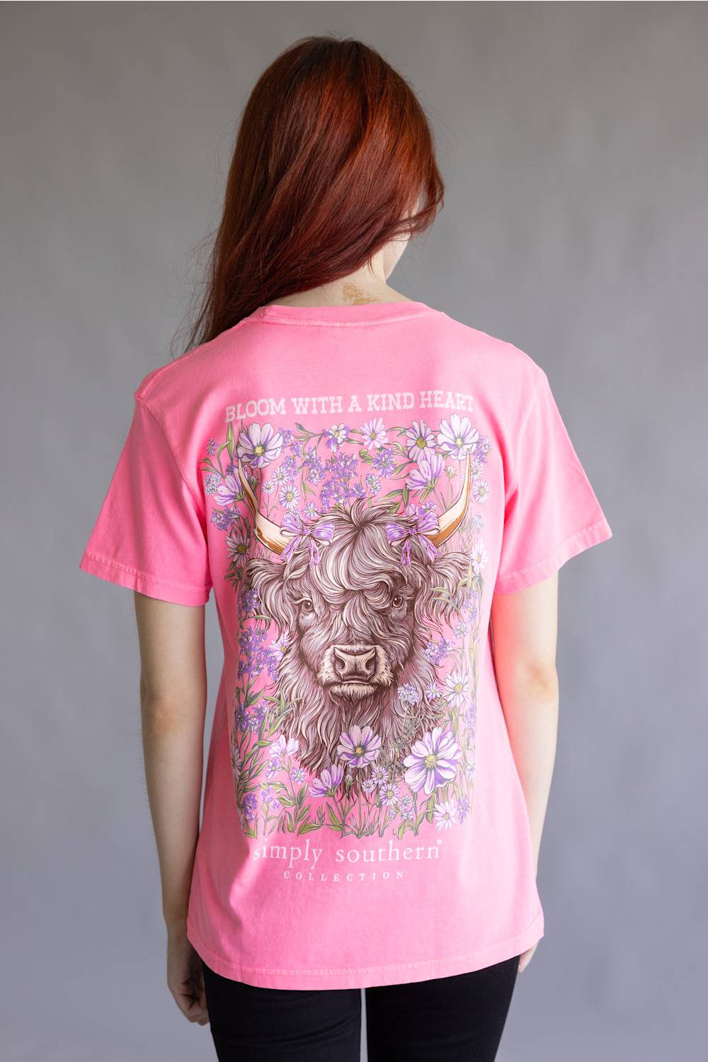 A woman viewed from behind wearing a bright pink graphic T-shirt. The shirt features a large illustration of a Scottish highland cow surrounded by purple and white flowers, with the text 'BLOOM WITH A KIND HEART' above and 'Simply Southern California' below.