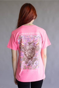 A woman viewed from behind wearing a bright pink graphic T-shirt. The shirt features a large illustration of a Scottish highland cow surrounded by purple and white flowers, with the text 'BLOOM WITH A KIND HEART' above and 'Simply Southern California' below.