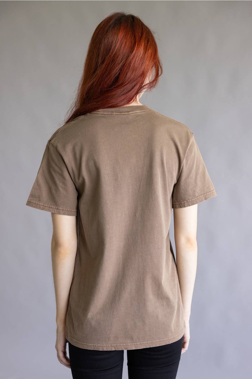 A person viewed from the back wearing a plain taupe-brown short-sleeve T-shirt and black pants, standing against a neutral gray studio background.