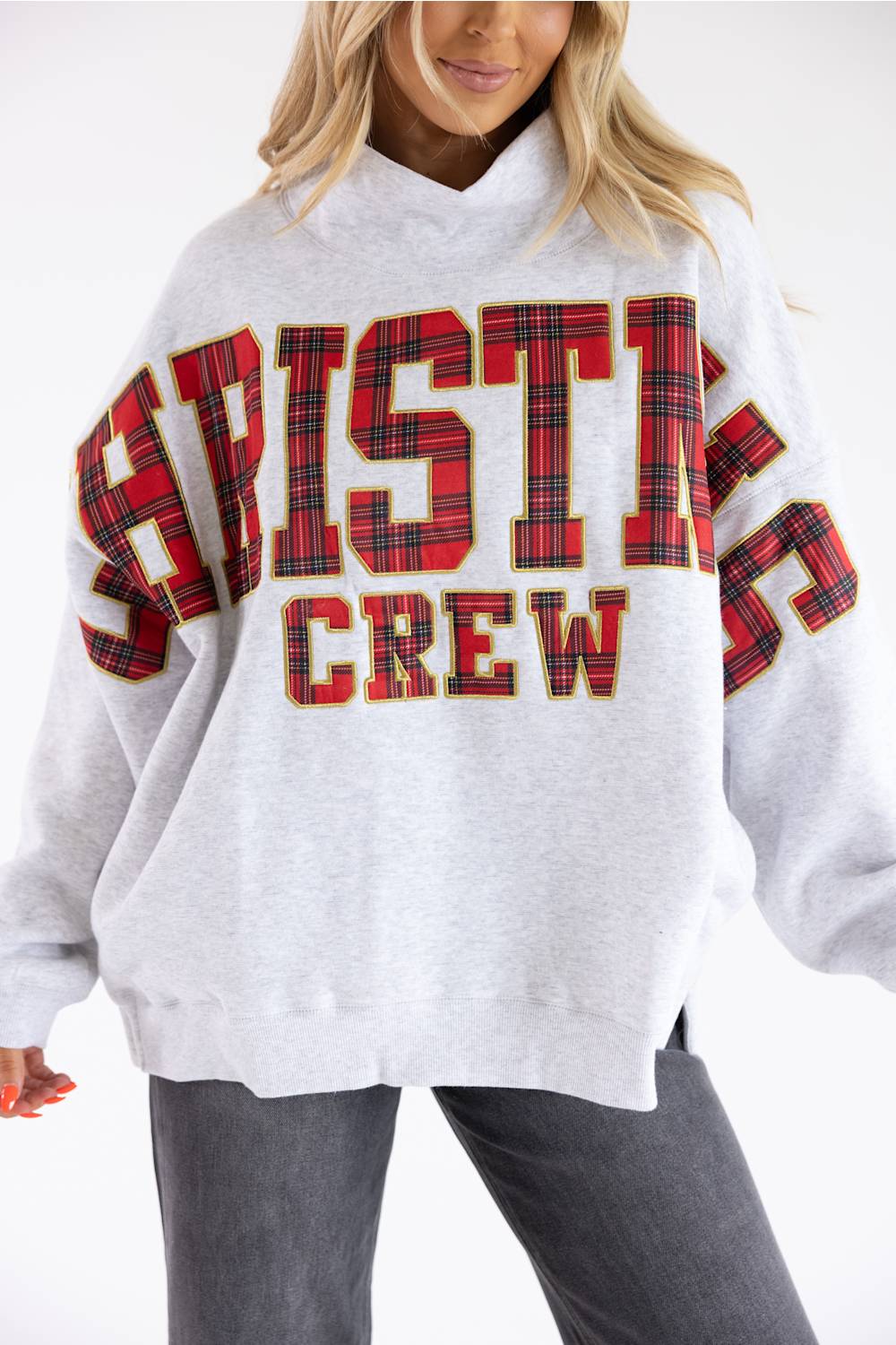 A woman wearing an oversized light gray sweatshirt with large red and black plaid collegiate-style lettering that reads 'CHRISTMAS CREW' across the front, paired with dark gray jeans against a plain white studio background.