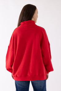 A woman viewed from the back wearing a bright red oversized mock-neck sweatshirt and blue jeans against a plain white studio background.