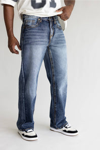 A person wearing loose-fit medium-wash blue denim jeans with a faded wash, paired with a white t-shirt and black-and-white sneakers, shown from the waist down against a light gray studio background.