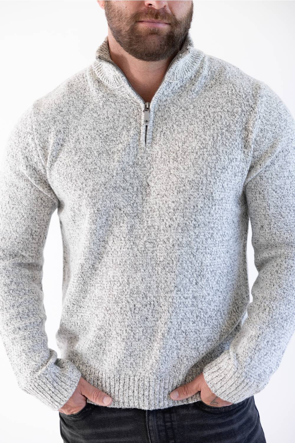 A man wearing a light gray and white heathered knit quarter-zip sweater with a stand collar, paired with dark jeans against a plain white background.