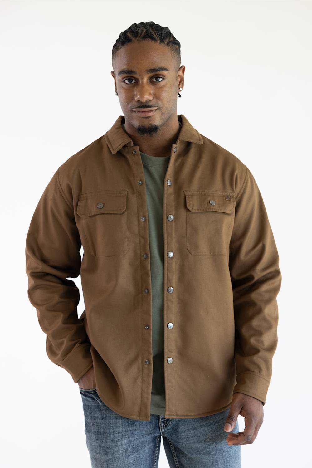 A man wearing a brown button-up overshirt over a green t-shirt and blue jeans, standing against a plain white studio background.