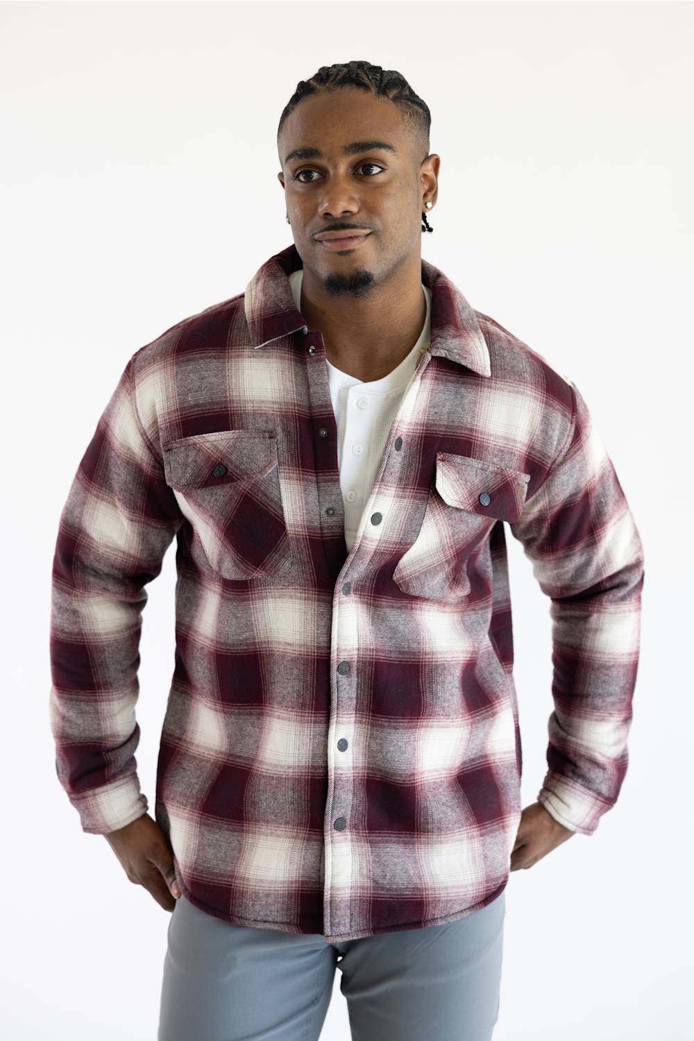 A man wearing a long-sleeve button-up plaid flannel shirt in burgundy and cream, layered over a white t-shirt and gray pants, standing against a plain white background.