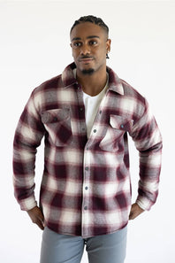 A man wearing a long-sleeve button-up plaid flannel shirt in burgundy and cream, layered over a white t-shirt and gray pants, standing against a plain white background.