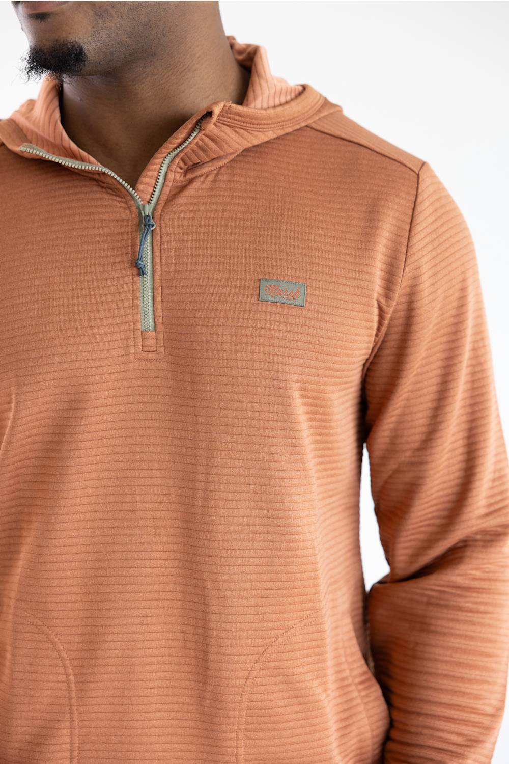 A close-up of a man wearing a textured, ribbed quarter-zip pullover in a warm terracotta or burnt orange color. The garment features a small rectangular logo patch on the left chest and a light-colored zipper pull.