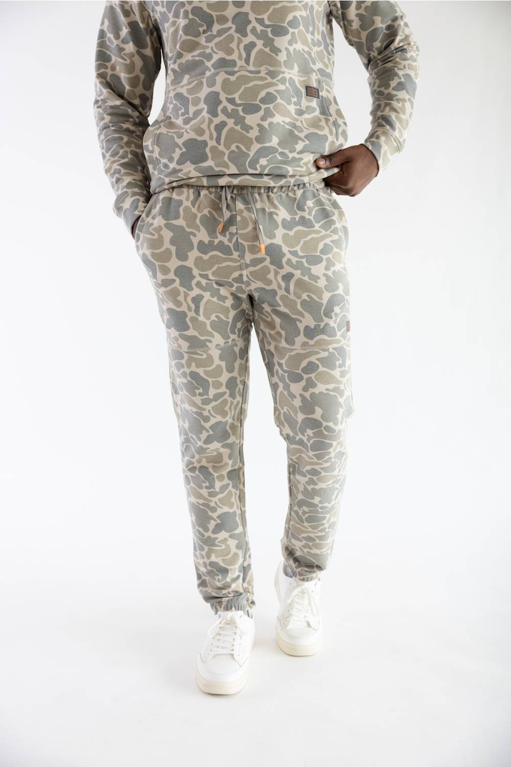 A person wearing a matching camouflage sweatshirt and jogger pants set in shades of beige, gray, and olive green, paired with white sneakers, photographed against a plain white studio background.